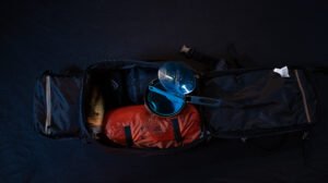 Vue aérienne d’un sac à dos de randonnée posé au sol, entouré d’équipements outdoor essentiels dispersés : gourde, vêtements techniques, réchaud, carte et trousse de secours.